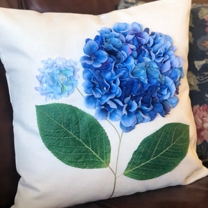 May include: A white pillow with a blue hydrangea flower design. The flower is made of fabric and has a green stem and leaves.