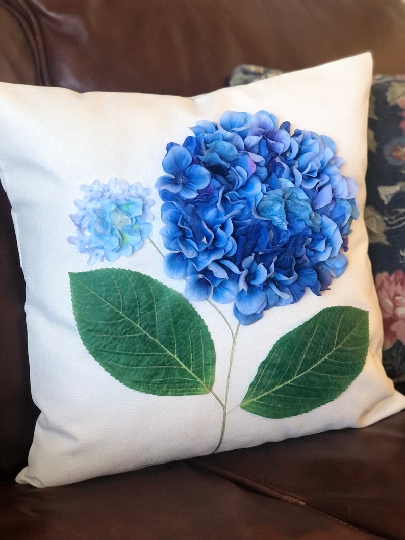 May include: A white pillow with a blue hydrangea flower design. The flower is made of fabric and has a green stem and leaves.