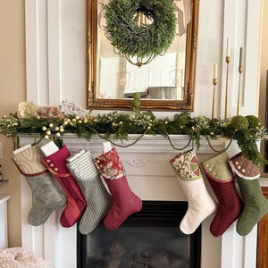 Könnte beinhalten: Sieben Weihnachtsstrümpfe hängen in einer Reihe, jeder mit einer einzigartigen Kombination aus Stoffen, Farben und Mustern, einschließlich einfarbiger, karierter und floraler Designs, akzentuiert mit Zierknöpfen an den oberen Bündchen.