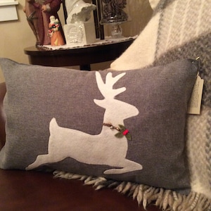 May include: A gray throw pillow with a white felt reindeer applique. The reindeer has a brown and red holly berry garland around its neck.