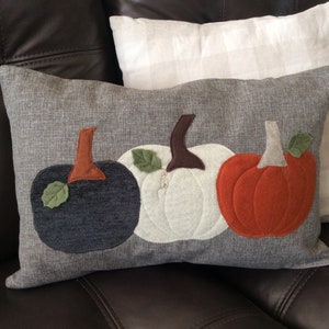 May include: Rectangular grey cushion with appliqued pumpkins in navy, white, and orange. Each pumpkin has a brown stem and a green leaf. A fall-themed decorative item for the home.
