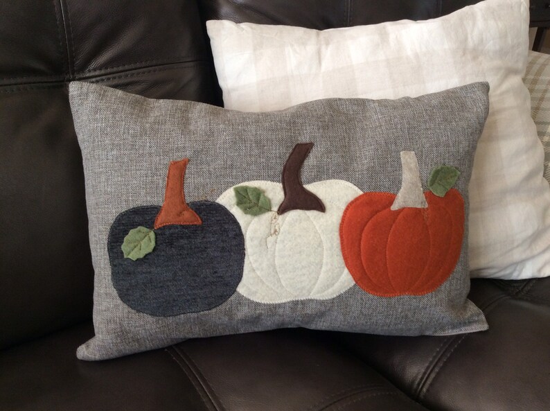 May include: Rectangular grey cushion with appliqued pumpkins in navy, white, and orange. Each pumpkin has a brown stem and a green leaf. A fall-themed decorative item for the home.