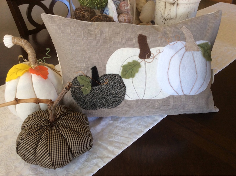 May include: A rectangular cushion featuring appliqu&eacute;d white and dark grey pumpkins. Two fabric pumpkins with stems and leaves are in front of the cushion. The pumpkins are white, and black and white checked.