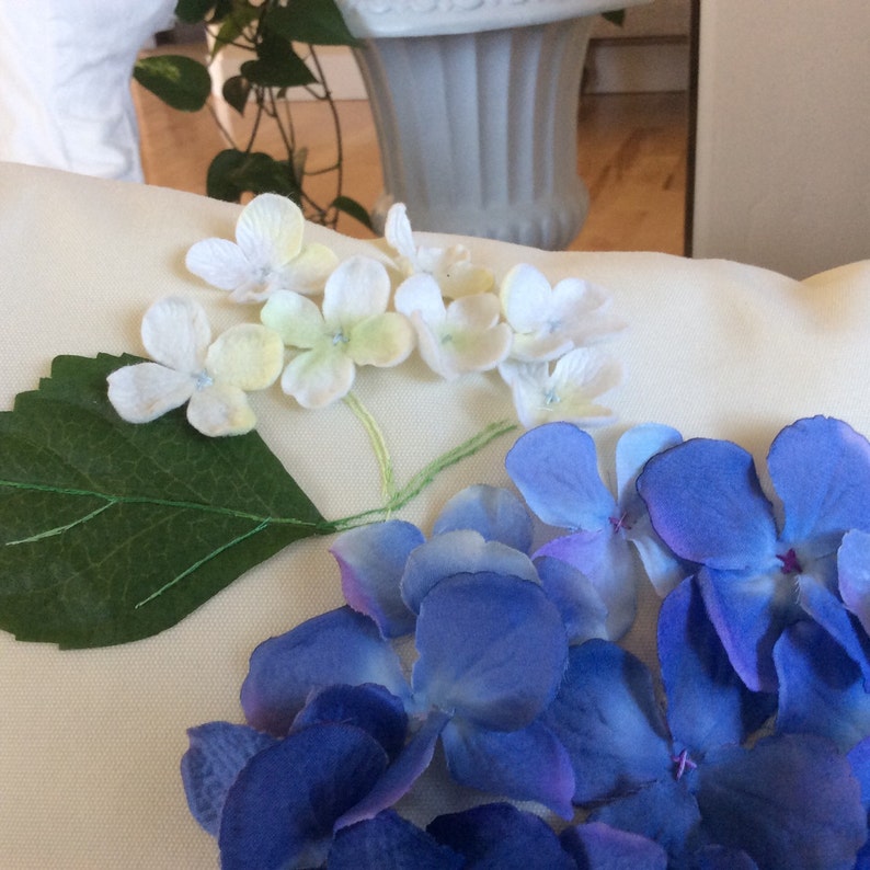 May include: A white pillow with a floral design. The pillow features white and yellow flowers with green leaves and blue flowers.