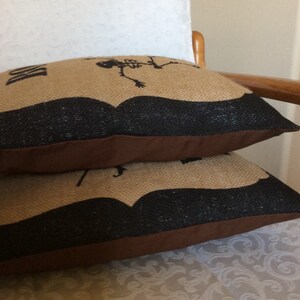 May include: Two decorative burlap pillows stacked on top of each other. The pillows have a tan background with a black mustache design and a skeleton graphic. The bottom edge of the pillows is brown.