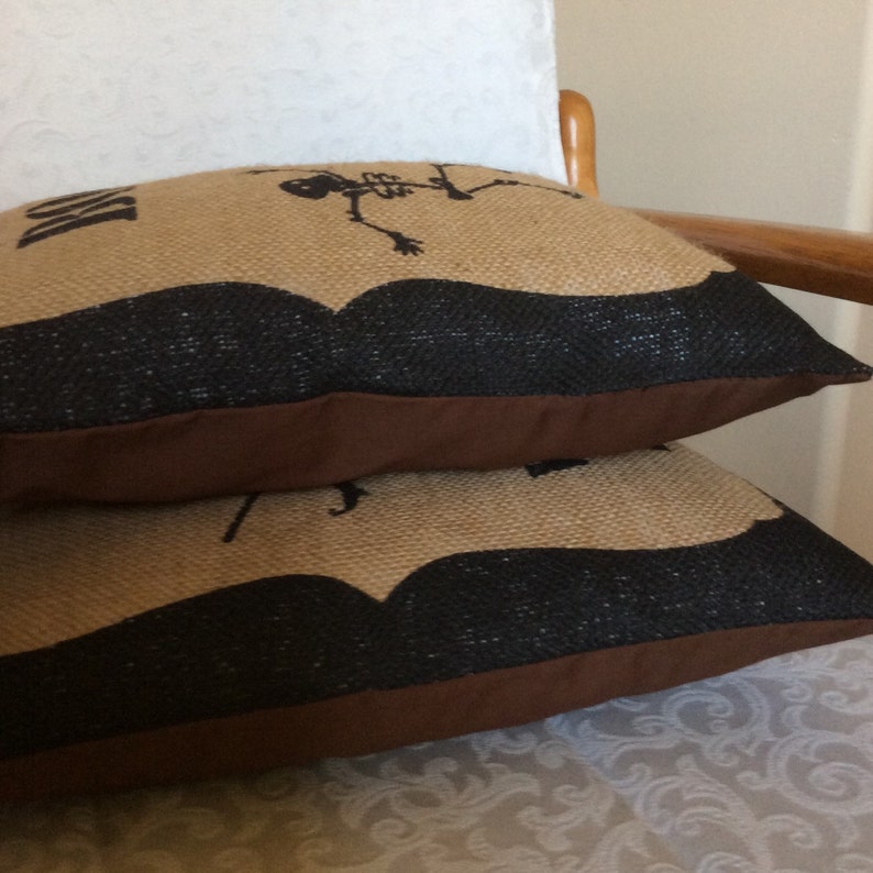 May include: Two decorative burlap pillows stacked on top of each other. The pillows have a tan background with a black mustache design and a skeleton graphic. The bottom edge of the pillows is brown.