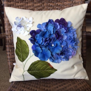May include: A white pillow with a blue and purple hydrangea flower embroidered on it. The flower is made of fabric petals and has green leaves.