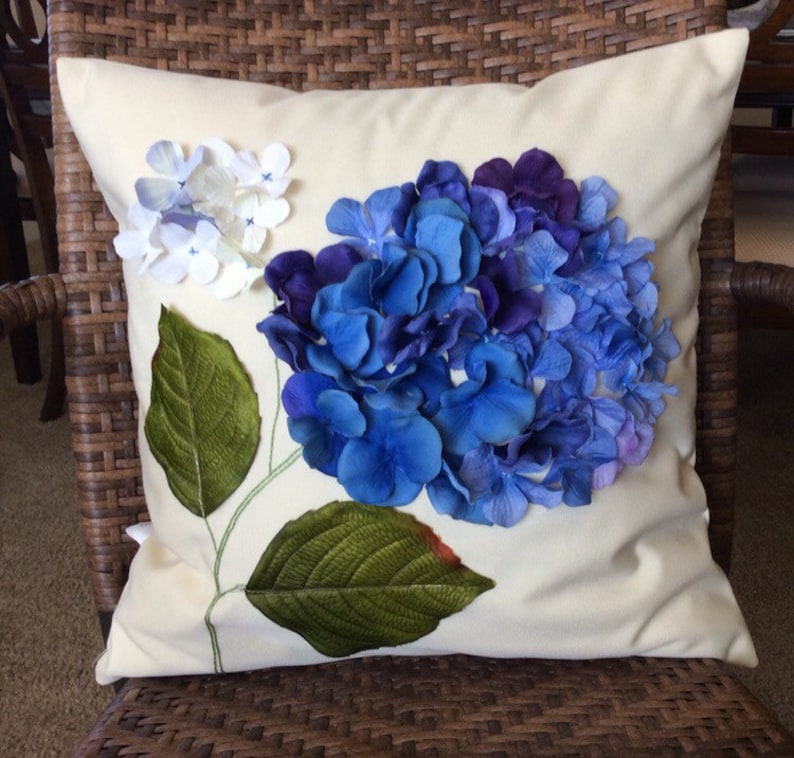 May include: A white pillow with a blue and purple hydrangea flower embroidered on it. The flower is made of fabric petals and has green leaves.