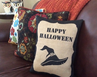 May include: A collection of decorative throw pillows on a brown leather couch. One pillow features the text "HAPPY HALLOWEEN" and a witch hat graphic. Other pillows have floral patterns and a solid orange pillow is in the background.
