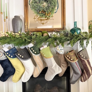 May include: A row of eight fabric Christmas stockings hanging from a mantel, each featuring a unique combination of colours, textures, and patterns, including solid, striped, and floral designs, all accented with small white buttons on the cuffs.