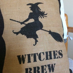 May include: A burlap pillow with a black silhouette of a witch on a broomstick. The words "WITCHES BREW" are printed in black block letters below the witch. The pillow has a natural, rustic appearance.