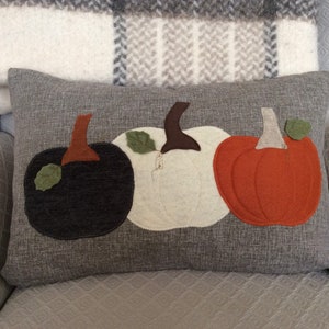 May include: A grey rectangular cushion featuring three felt pumpkins in black, white, and orange. Each pumpkin has a brown stalk and a green leaf. The cushion is on a light grey chair with a plaid blanket.