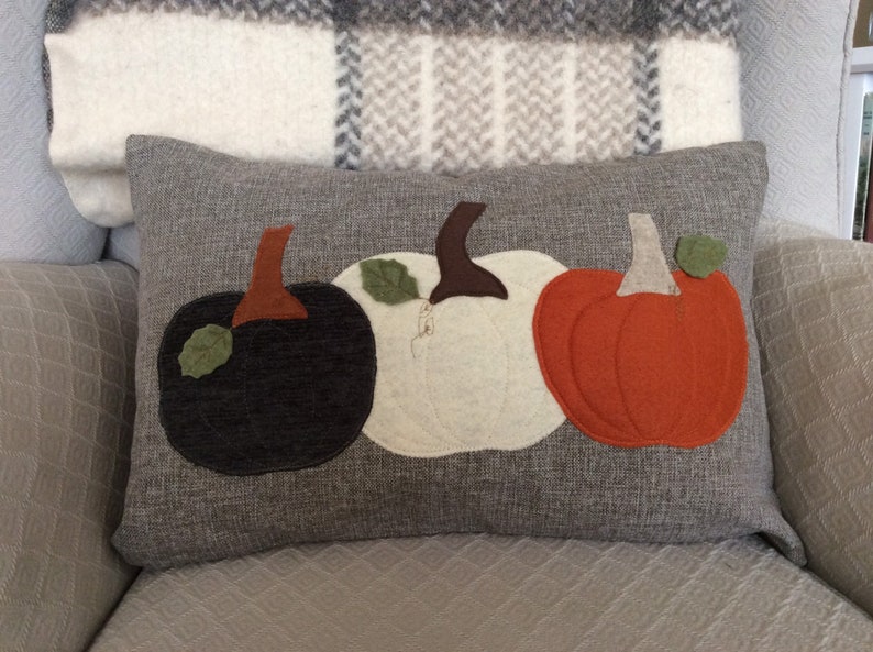 May include: A grey rectangular cushion featuring three felt pumpkins in black, white, and orange. Each pumpkin has a brown stalk and a green leaf. The cushion is on a light grey chair with a plaid blanket.