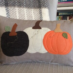 May include: A rectangular cushion with three felt pumpkins in black, white, and orange. Each pumpkin has a brown stem and a green leaf. The cushion is on a grey background, suggesting an autumn or harvest theme.