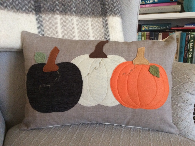 May include: A rectangular cushion with three felt pumpkins in black, white, and orange. Each pumpkin has a brown stem and a green leaf. The cushion is on a grey background, suggesting an autumn or harvest theme.