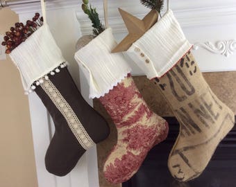 May include: Three Christmas stockings hang from a mantel. One is brown with a cream lace stripe and white trim. The second is patterned with red and cream floral designs. The third is made of burlap with printed text.