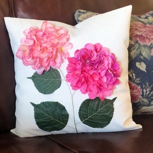 May include: White square decorative pillow featuring two large pink hydrangea flower appliqués with green leaves and stems. The pillow is on a brown leather surface.