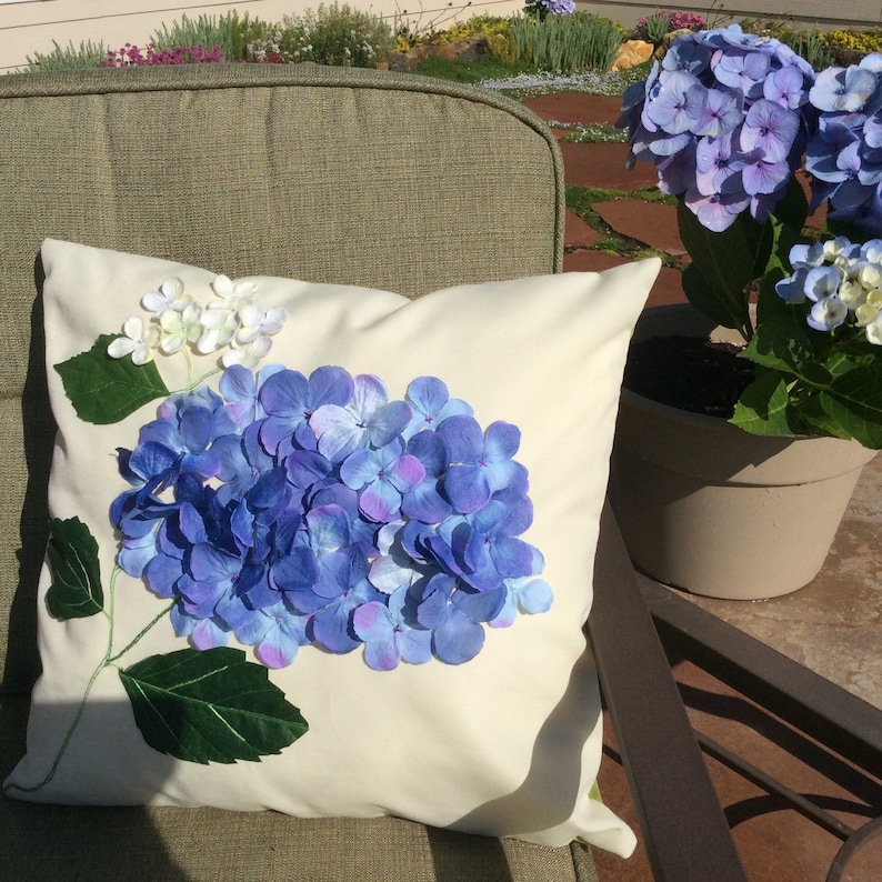 May include: A white pillow with a blue and purple hydrangea flower design. The pillow is on a green chair.