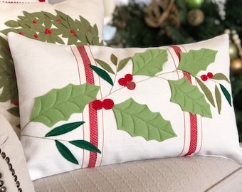 May include: Rectangular white pillow with a holly and berry design. The pillow features green felt holly leaves and red berries, with red and white striped accents. Other pillows with holiday designs are visible.