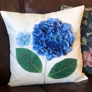 May include: White throw pillow with a blue hydrangea flower printed on it. The flower is in the center of the pillow and has green leaves.