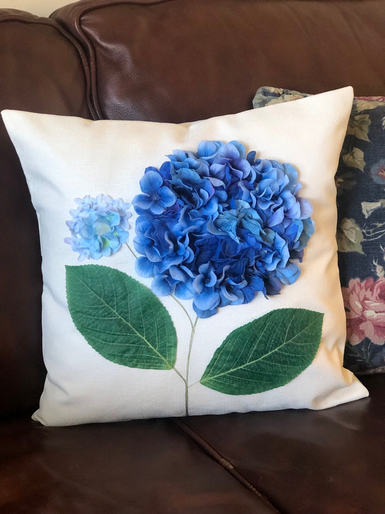 May include: White throw pillow with a blue hydrangea flower printed on it. The flower is in the center of the pillow and has green leaves.