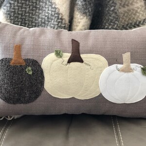 May include: A rectangular grey decorative cushion featuring three appliqu&eacute;d pumpkins in black, cream, and white. Each pumpkin has a brown stem and a green leaf. The cushion is on a grey surface.