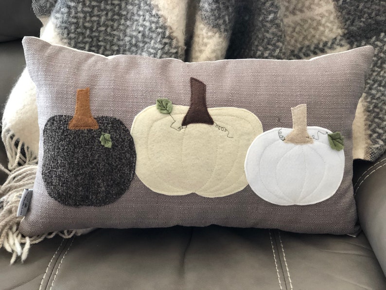 May include: A rectangular grey decorative cushion featuring three appliqu&eacute;d pumpkins in black, cream, and white. Each pumpkin has a brown stem and a green leaf. The cushion is on a grey surface.