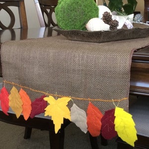 May include: A brown burlap table runner with a string of felt fall leaves in shades of red, orange, yellow, and green.