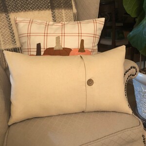 May include: Cream-coloured rectangular cushion with a large wooden button closure. Behind it, a cushion with a white plaid pattern and pumpkin appliqu&eacute;s. The cushions are on a patterned armchair, with a plaid blanket in the background.