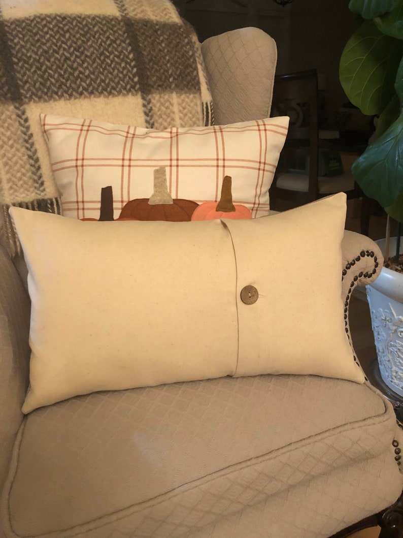 May include: Cream-coloured rectangular cushion with a large wooden button closure. Behind it, a cushion with a white plaid pattern and pumpkin appliqu&eacute;s. The cushions are on a patterned armchair, with a plaid blanket in the background.