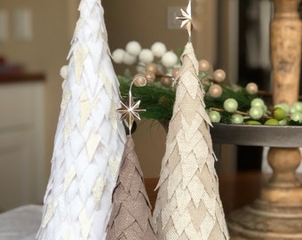 May include: Three decorative Christmas trees in varying sizes and neutral tones. The trees are covered in fabric with a diamond pattern. Each tree is topped with a gold star. The largest tree is white, the medium tree is beige, and the smallest tree is brown.