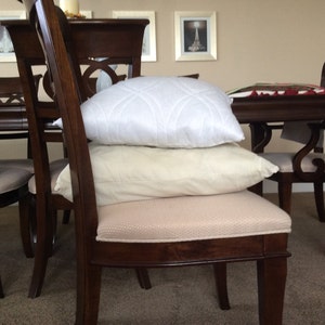 May include: A set of four wooden dining chairs with padded seats. Two chairs have white and cream pillows on the seats. The chairs are arranged around a wooden table.