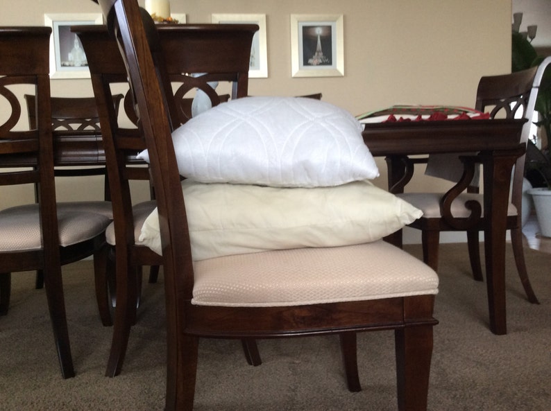 May include: A set of four wooden dining chairs with padded seats. Two chairs have white and cream pillows on the seats. The chairs are arranged around a wooden table.