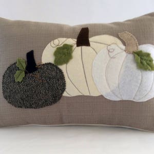 May include: A rectangular cushion featuring three appliqu&eacute;d pumpkins. The pumpkins are in shades of grey and white, with green leaves and brown stalks. The cushion has a neutral, textured background.