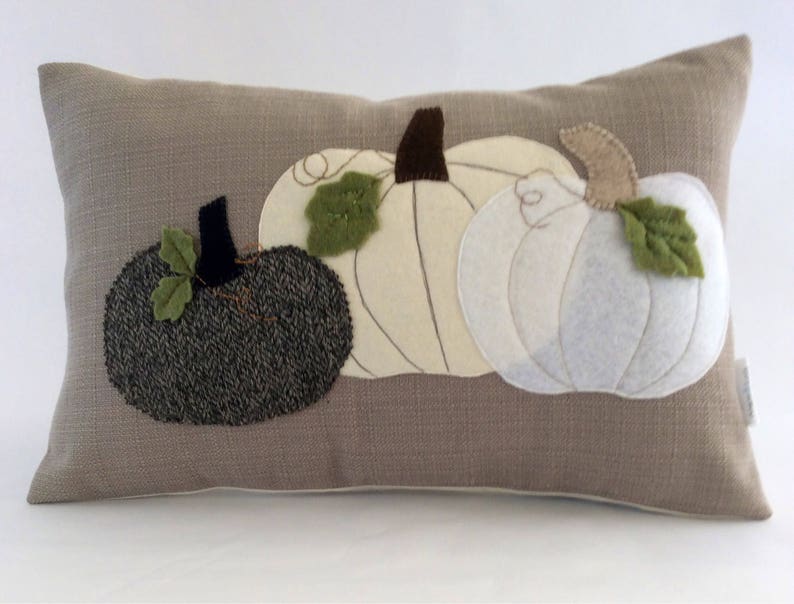 May include: A rectangular cushion featuring three appliqu&eacute;d pumpkins. The pumpkins are in shades of grey and white, with green leaves and brown stalks. The cushion has a neutral, textured background.