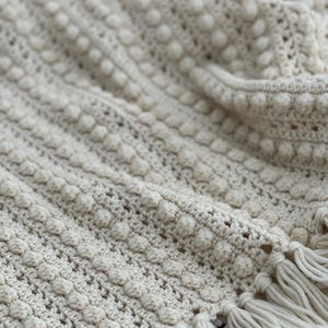 May include: Cream-colored, hand-crocheted blanket with a textured, bobble stitch pattern. The blanket features a fringe along the bottom edge. The soft, neutral color and detailed stitching create a cozy, handmade aesthetic.