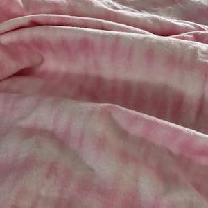 May include: Pink and white tie-dye fabric with soft folds and a textured appearance. The fabric has a watercolor effect, with the pink dye bleeding into the white background. The material appears to be soft and lightweight.