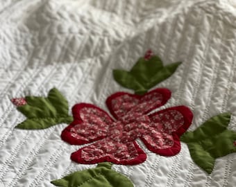 May include: Close-up of a white quilt with a floral design. The quilt features red and white floral appliqués with green leaves. The quilt has a textured, quilted surface with a diamond pattern.