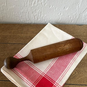 May include: A vintage wooden muddler with a rounded handle rests on a red and white checkered linen napkin. The muddler is a dark brown color and appears to be well-used. The napkin is folded and placed on a wooden surface.