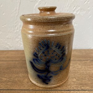 May include: A brown ceramic jar with a lid, featuring a blue and white floral design. The jar has a rustic, handmade look.