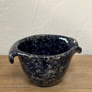 May include: A speckled blue ceramic bowl with a curved handle on each side. The bowl has a dark blue base color with lighter speckles throughout. The bowl is sitting on a wooden surface.