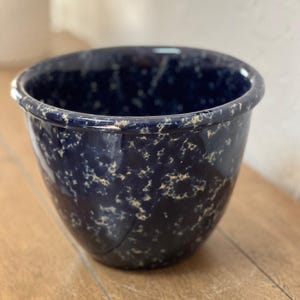 May include: A navy blue ceramic bowl with a speckled pattern of cream-colored spots. The bowl has a rounded shape with a slightly flared rim. The bowl is sitting on a wooden surface.