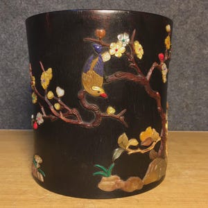 May include: A dark brown wooden cylindrical container with carved relief details. The relief depicts a bird perched on a flowering branch with yellow, white, and red blossoms. The container is set on a light brown surface.