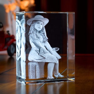 May include: A 3D crystal cube with a laser-engraved image of a young girl wearing a hat and a dress, sitting on a suitcase. The image is in black and white.