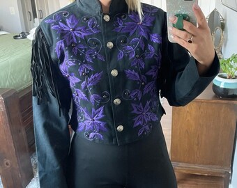 May include: A black suede jacket with purple floral embroidery and fringe detailing. The jacket has a button-up front and a cropped style.