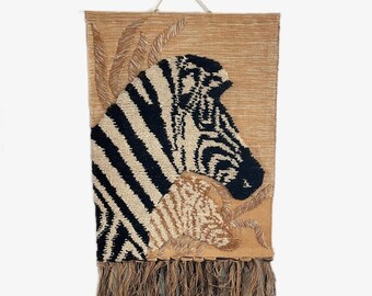 May include: A woven wall hanging featuring a zebra head with black and white stripes and a tan background. The artwork includes a fringe at the bottom and decorative elements. The piece is designed to be hung on a wall.