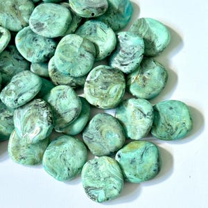 May include: A collection of turquoise-colored, irregular-shaped beads with a marbled effect. The beads have a flat, disc-like shape and are scattered across a white surface. The beads have a hole for stringing.