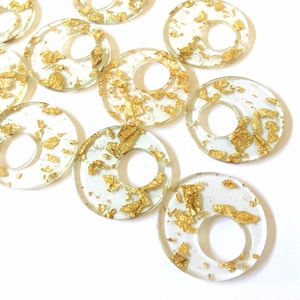 May include: A group of clear resin rings with gold flakes embedded inside. The rings have a hole in the center and are scattered on a white background.