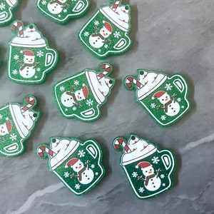 May include: Green glittery Christmas-themed mug-shaped embellishments. Each mug features a snowman wearing a red hat and scarf, snowflakes, and a candy cane. The mugs have white whipped cream and a candy cane straw.