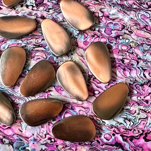 May include: A collection of ten smooth, oval-shaped copper-colored metal pieces. The pieces are arranged on a pink, blue, and white iridescent background.